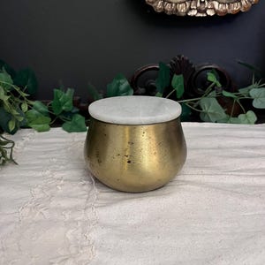May include: A small, antique brass jar with a white marble lid. The jar has a rounded shape and a slightly aged appearance. The lid sits flush on top. The jar is displayed on a white lace cloth, suggesting a vintage or decorative theme.