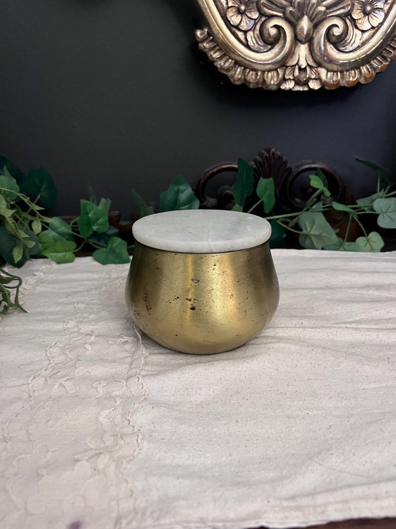 May include: A small, antique brass jar with a white marble lid. The jar has a rounded shape and a slightly aged appearance. The lid sits flush on top. The jar is displayed on a white lace cloth, suggesting a vintage or decorative theme.