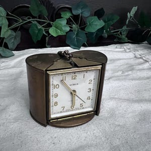 May include: A vintage brass travel clock with a rectangular face and a hinged cover. The clock face is white with black numerals and hands, and the brand name "semca" is visible. The clock is set against a neutral background.