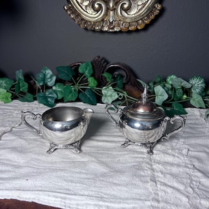 Peut inclure: Un ensemble de crémier et sucrier en argent. Le crémier a une poignée incurvée et repose sur trois petits pieds. Le sucrier a un couvercle et des pieds assortis. Les deux pièces sont très réfléchissantes.