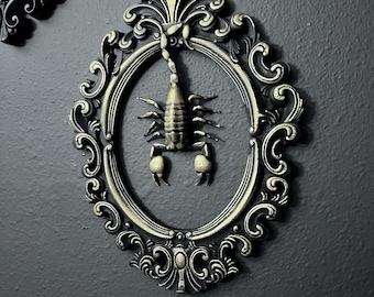 May include: Ornate, oval-shaped wall decor featuring a detailed scorpion within a decorative frame. The frame has a dark, antique gold finish with intricate scrollwork. The scorpion is a metallic gold color, set against a dark gray wall.