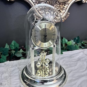 Puede incluir: Un reloj de aniversario plateado bajo una cúpula de cristal transparente. El reloj presenta una esfera redonda con números romanos negros, manecillas negras ornamentadas y la palabra "Comitti" impresa. Se asienta sobre una base circular plateada.