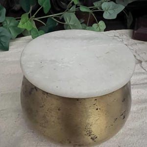 May include: A round, brass-colored container with a white, circular lid. The container has a slightly textured surface. The lid appears to be made of marble or a similar stone. The overall aesthetic is vintage or antique.