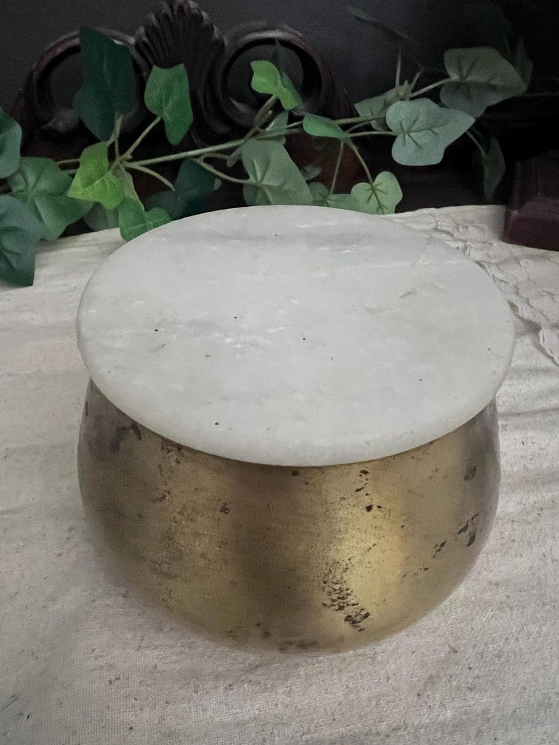 May include: A round, brass-colored container with a white, circular lid. The container has a slightly textured surface. The lid appears to be made of marble or a similar stone. The overall aesthetic is vintage or antique.