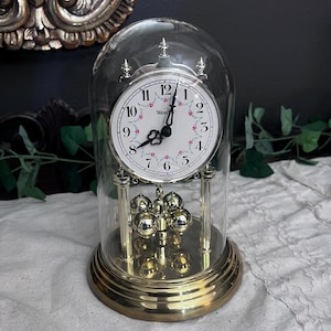 May include: A brass-toned clock with a white face, floral accents, and a glass dome. The clock has a pendulum with multiple brass spheres. The clock face has black Roman numerals and black hands. The clock is a decorative item.