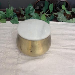 May include: A small, round brass container with a white marble lid. The container has a slightly tarnished, aged appearance. The lid is a smooth, circular piece of white marble. The container is sitting on a white, textured surface.