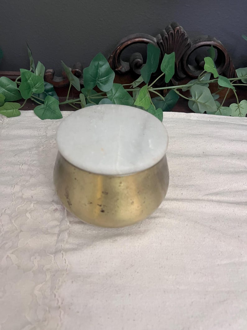 May include: A small, round brass container with a white marble lid. The container has a slightly tarnished, aged appearance. The lid is a smooth, circular piece of white marble. The container is sitting on a white, textured surface.
