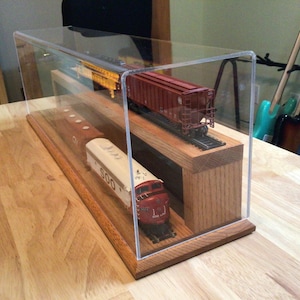May include: A clear acrylic display case with a wooden base containing two model trains on tracks. The top level features a red freight car, while the bottom level holds a red and white locomotive with the text SOO printed on the side.