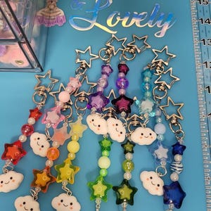May include: A collection of colorful keychains with star-shaped clasps. Each keychain features a string of beads, star charms, and smiling cloud charms in a rainbow of colors. The text "Customized & Lovely" is visible in the background.