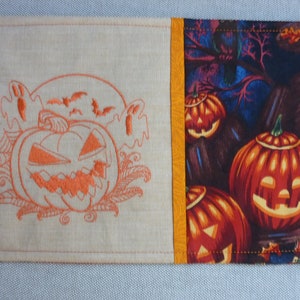 Könnte beinhalten: Ein Halloween-Platzset mit einem orangefarbenen, gestickten Kürbis, Fledermäusen und einem Geist auf beigem Hintergrund. Die rechte Seite zeigt ein Muster aus geschnitzten Kürbissen und eine dunkle Waldszene.