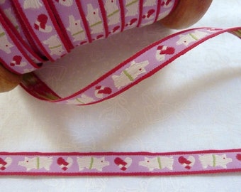 May include: A spool of decorative ribbon with a repeating pattern of white dogs, red hearts, and green accents on a lavender background. The ribbon has a red border and is ideal for crafts and gift wrapping.