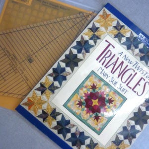 Puede incluir: Un libro titulado "Triangles: A New Twist on Triangles" de Mary Sue Suit, con un diseño de edredón colorido en la portada. Una regla triangular de plástico transparente es visible, ideal para proyectos de acolchado.