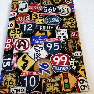 May include: A fabric item with a black background covered in colorful road signs. The signs include Route 66, Interstate, speed limit, and other road-related graphics. The item is likely a decorative piece or fabric.