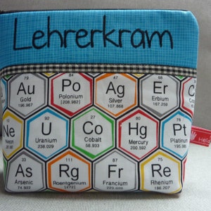 Puede incluir: Una bolsa azul con cremallera con el texto "Lehrerkram" bordado en la parte delantera. La bolsa está decorada con un patrón de hexágonos coloridos, cada uno con el símbolo y el nombre de un elemento químico de la tabla periódica.