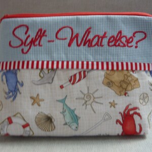 Peut inclure: Une pochette zippée blanche avec un motif vichy bleu et blanc et une bordure rouge. La pochette est ornée d'un texte brodé en rouge qui dit "Sylt - What else?" et d'une variété d'illustrations sur le thème nautique, notamment un crabe, une étoile de mer, une coquille et un poisson.