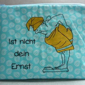Könnte beinhalten: Eine türkisfarbene Reißverschlusstasche mit einem sich wiederholenden weißen Wirbelmuster. Die Tasche zeigt eine gestickte Cartoonfigur in gelber Kleidung, die auf einer Waage steht. Der Text "Ist nicht dein Ernst" ist schwarz gestickt.