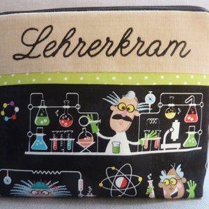Può includere: Una pochette con cerniera nera e verde con uno sfondo bianco e il testo "Lehrerkram" ricamato in alto. La pochette presenta un'illustrazione in bianco e nero di uno scienziato in un laboratorio con becher, provette e altre attrezzature scientifiche.