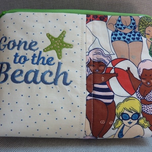 Peut inclure: Une pochette en tissu blanc à pois bleus avec le texte "Gone to the Beach" brodé en bleu. La pochette a une fermeture éclair verte et une doublure en tissu coloré sur le thème de la plage avec des femmes en maillot de bain et un ballon de plage.