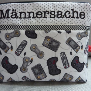 May include: A white zippered pouch with a black and white chevron pattern and the text "Männersache" embroidered on the front. The pouch is decorated with a repeating pattern of black and grey video game controllers and handheld game consoles.