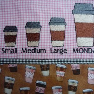 May include: Embroidered design on pink and white gingham fabric featuring four coffee cups of different sizes labeled "Small", "Medium", "Large", and "MONDAY". The bottom of the image shows a brown fabric with a repeating pattern of coffee cups.