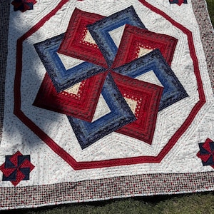 May include: A handmade quilt with a starburst design in red, white, and blue. The quilt features a central octagonal pattern with alternating red and blue squares, surrounded by a white background with floral accents. The border is a red and white gingham pattern.