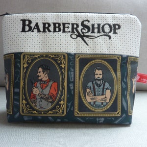 Puede incluir: Un neceser rectangular con cremallera negra, con un diseño de "BARBER SHOP" con tijeras. La bolsa muestra retratos de barberos en marcos ornamentados, sobre un fondo verde oscuro con herramientas de barbero.