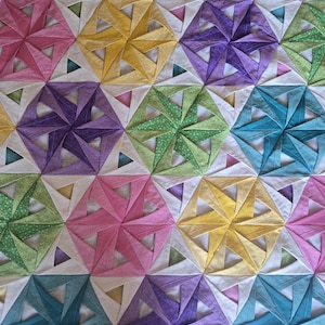 以下が含まれることがあります： 幾何学模様のカラフルなキルト。ピンク、黄色、紫、緑、青など、さまざまな色の六芒星が繰り返されるデザインです。星は白い背景に配置されています。