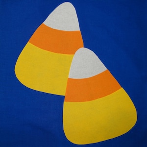 May include: Two felt candy corn shapes on a blue background. The candy corn is yellow, orange, and white.