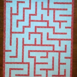 May include: A white fabric maze with a red border. The maze is made up of a grid of squares with paths cut out of them. The paths are all the same width and are all straight lines.