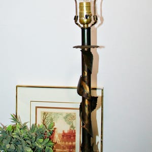 Może przedstawiać: Ozdobna lampa stołowa z brązowym wykończeniem. Lampa posiada dekoracyjny, skręcony trzon z akcentami w kształcie liści i przezroczystą żarówkę w kształcie łzy. Lampa stoi na czarnej podstawie.