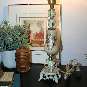 Puede incluir: Una lámpara de mesa vintage con una pantalla de cristal crema decorada con flores y una base de latón. La lámpara tiene una base decorativa y ornamentada y una bombilla clara y puntiaguda. La lámpara está sobre una superficie negra, junto a un cuadro enmarcado y una planta en maceta.