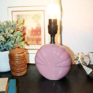 May include: A vintage pink ceramic table lamp with a decorative fan design. The lamp has a black base and a clear, teardrop-shaped lightbulb. A framed print and a wooden container are in the background.