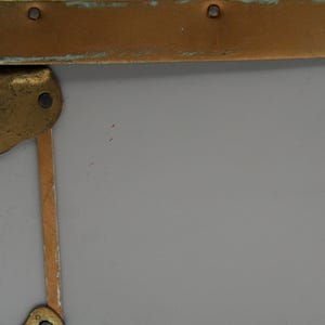 May include: Close-up of a gray trunk corner with brass hardware. The trunk has brass corner protectors and a brass band along the top edge. The hardware shows signs of age and wear.