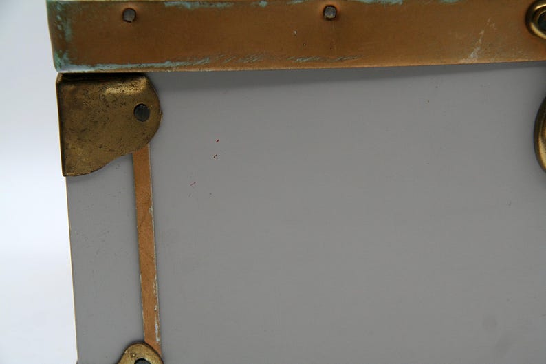 May include: Close-up of a gray trunk corner with brass hardware. The trunk has brass corner protectors and a brass band along the top edge. The hardware shows signs of age and wear.