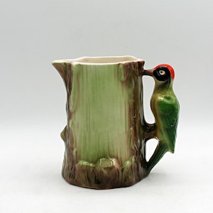 May include: A ceramic pitcher shaped like a tree trunk with a light green center and brown bark texture at the base and rim. A green woodpecker with a bright red crown and black facial markings serves as the handle, attached to the side of the trunk.