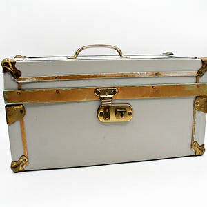May include: A vintage-style gray trunk with brass accents. The trunk features a brass handle, corner protectors, and a central clasp. The metal trim and hardware provide a classic aesthetic. The trunk is a rectangular shape and sits on a white background.