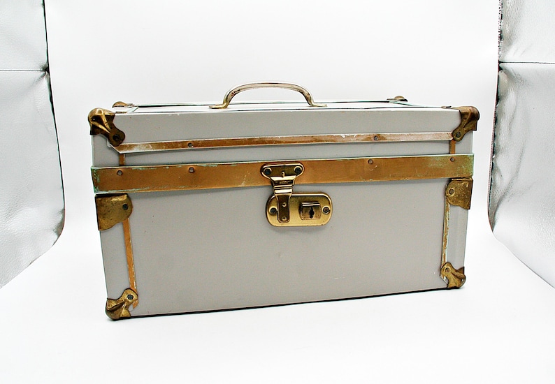 May include: A vintage-style gray trunk with brass accents. The trunk features a brass handle, corner protectors, and a central clasp. The metal trim and hardware provide a classic aesthetic. The trunk is a rectangular shape and sits on a white background.