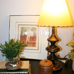 May include: A table lamp with a cream-coloured, scalloped lampshade. The lamp sits on a dark surface next to a framed print of a European town, a small plant, a wooden jar, and a stack of books. The lamp has a brass base.