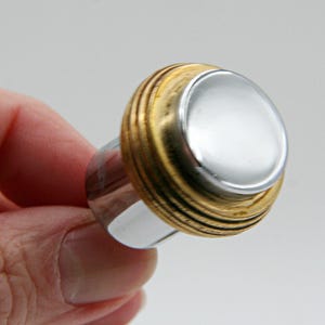 May include: A cylindrical bottle stopper featuring a polished silver-tone top disc, a central band with three horizontal gold-tone ridges, and a smooth, reflective silver-tone base that tapers slightly toward the bottom end.