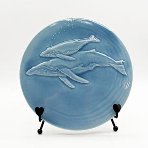 May include: A round, light blue ceramic plate featuring a raised, embossed design of two whales swimming together. The larger whale is positioned below a smaller whale, with small bubbles rising above them. The plate rests on a black metal stand.