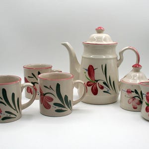 May include: A cream-colored ceramic tea set with a floral design. The set includes a teapot, sugar bowl, creamer, and four mugs. Each piece is decorated with red flowers and green leaves.