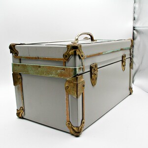 May include: A vintage gray trunk with brass-colored corner protectors, latches, and handle. The trunk has a rectangular shape and is likely used for storage or travel. The metal accents show signs of age and wear, adding to its antique charm.