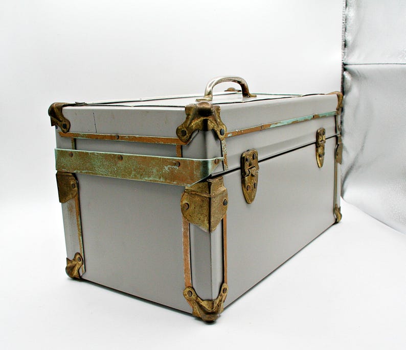 May include: A vintage gray trunk with brass-colored corner protectors, latches, and handle. The trunk has a rectangular shape and is likely used for storage or travel. The metal accents show signs of age and wear, adding to its antique charm.