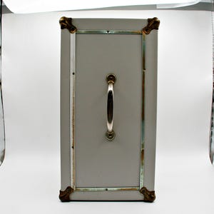 May include: A rectangular, gray storage trunk with a silver handle and brass-colored corner protectors. The trunk has a rectangular door with a silver frame and is likely vintage or antique.