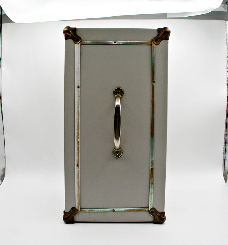 May include: A rectangular, gray storage trunk with a silver handle and brass-colored corner protectors. The trunk has a rectangular door with a silver frame and is likely vintage or antique.
