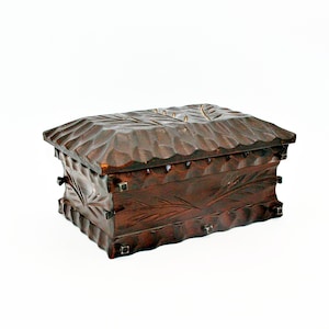 May include: A rectangular dark brown wooden box with a hinged lid. The exterior features a hand-carved texture with scalloped edges and engraved leaf patterns on the sides and top. Small metal square accents are visible at the corners.
