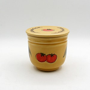 May include: A yellow ceramic jar with a matching lid, featuring hand-painted red tomato illustrations on the front and a small tomato graphic surrounded by a brown leaf wreath design on the top of the lid.