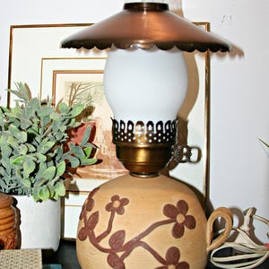 May include: A table lamp with a tan ceramic base featuring brown floral relief patterns and a side handle. It has a brass-toned metal collar, a white glass globe shade, and a wide, scalloped-edge copper-colored metal shade topper with a clear chimney.