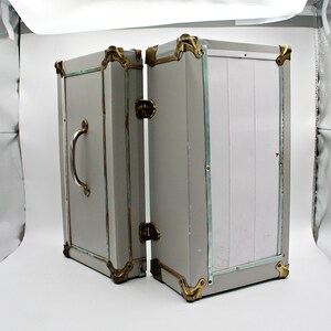May include: A vintage-style gray trunk with brass-colored corner protectors and accents. The trunk is open, revealing the interior. It has a silver handle and a latch. The trunk is likely used for storage or decoration.