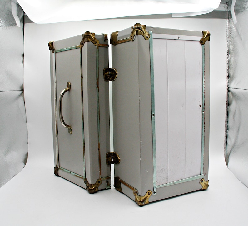 May include: A vintage-style gray trunk with brass-colored corner protectors and accents. The trunk is open, revealing the interior. It has a silver handle and a latch. The trunk is likely used for storage or decoration.